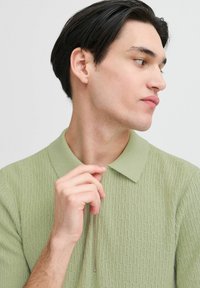 Lys grønn strikket polo skjorte med et teksturert mønster, knappeliv, og glidelås. Modellen har kort mørkt hår og vises fra siden.