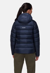 Marine pufferjack met een capuchon, met horizontaal gequiltde secties en een logo op de achterkant. Het materiaal lijkt glanzend en warm.