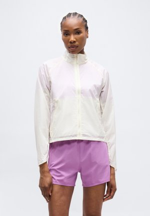 Žena s pletenými vlasmi, oblečená v ľahkej bielej bunde na zips s dlhými rukávmi a fialových športových šortkách, stojaca pred bielym pozadím.