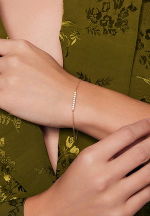 Złota bransoletka z delikatnym łańcuszkiem oraz rzędem małych, przejrzystych kamieni szlachetnych, noszona na ręce na tle oliwkowo-zielonej, teksturowanej tkaniny.