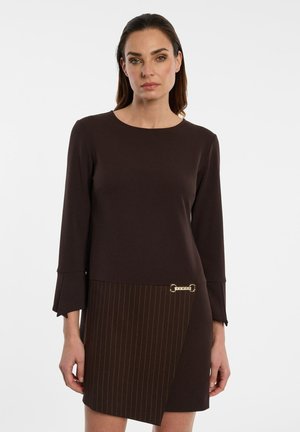 Vrouw draagt een donkerbruine jurk met lange mouwen met een gestreept rokgedeelte en goudkleurige kettingdetail, staand tegen een effen witte achtergrond.