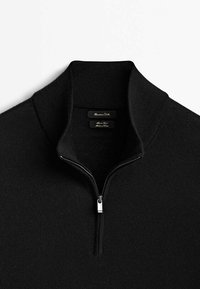 Pull noir à col zippé en tissu maille fine. Il présente un col montant, une texture lisse et un détail de fermeture éclair métallique au niveau du cou.