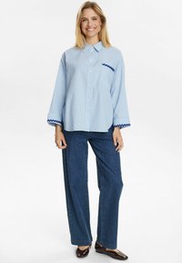 Ljust blå rutigt skjorta med knäppning, med mörkblå zigzagdetaljer på ärmsluten och bröstfickan. Matchas med mörkblå vida jeans.