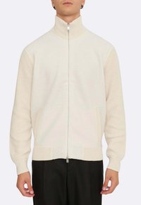 Cremefarbener Reißverschluss-Pullover mit hohem Kragen, gerippten Akzenten an Ärmeln und Saum, zwei seitlichen Taschen und einer glatten Stoffstruktur.