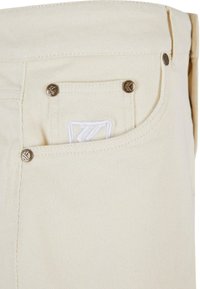 Pantaloni in corduroy beige con una finitura testurizzata, caratterizzati da bottoni bronzei e un logo ricamato bianco sulla piccola tasca frontale.
