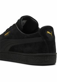 Sneaker nera in suede con suola in gomma nera testurizzata. Presenta il logo Puma dorato sul lato e sul retro, design elegante con accenti minimalisti.