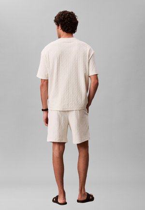 Mand stående med ryggen til iført en hvid tekstureret kortærmet skjorte og matchende shorts med sorte sandaler og en sort armbåndsur.