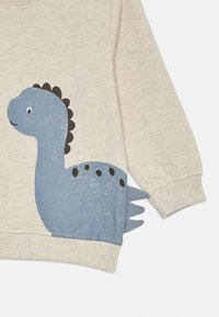 Barntröja i beige, med en blå dinosaurie-grafik med svarta fläckar och fjäll, tillverkad av mjukt, texturerat tyg.