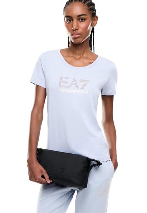 Giovane donna con capelli intrecciati indossa una t-shirt azzurro chiaro EA7 Emporio Armani e pantaloni, tenendo una pochette nera con il logo EA7.