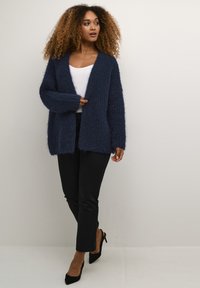 Tumši zils adīts kardigans ar atvērtu priekšpusi, garām piedurknēm, teksturētu audumu, valkāts pār baltu topu un apvienots ar melniem biksēm un augstpapēžu kurpēm.