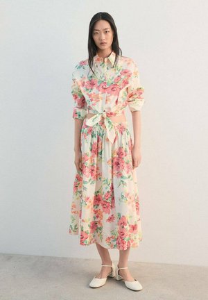 Robe longue fleurie crème avec motifs verts et roses, dotée d'un col à boutons et d'une taille nouée. Complétée par des chaussures blanches à talons blocs.