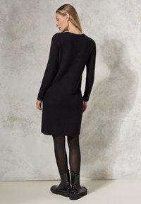 Schwarzes, geripptes Strickkleid mit langen Ärmeln und gerader Silhouette, kombiniert mit schwarzen Knöchelstiefeln. Glatte Textur und minimales Design.