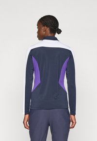 Langermet treningsgenser i marineblå med hvite ermer og lilla detaljer. Har en høy krage og et tettsittende design passende for trening.