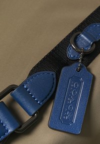 Étiquette en cuir bleu avec texte en relief, attachée à une sangle tissée noire, sur un fond vert olive mat. Design élégant et moderne.