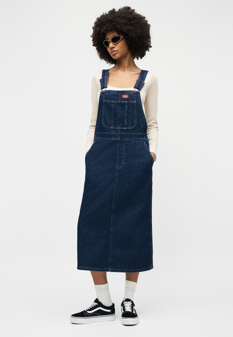 Frau mit Afro-Haaren, die eine Sonnenbrille, ein langes Jeans-Gesamtkleid, ein cremefarbenes Langarmoberteil, weiße Socken und schwarze Sneakers trägt und mit den Händen in den Taschen steht.