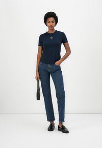 T-shirt bleu marine à manches courtes avec un col rond, associé à un jean bleu taille haute et des mocassins noirs, tenant un petit sac noir.