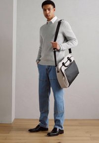 Grauer Pullover, weißes Hemd mit Kragen und blaue Jeans. Schwarz-graue Umhängetasche mit zwei Griffen und einem Schulterriemen. Schwarze Schuhe.