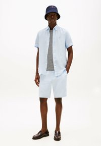 Camicetta a maniche corte azzurra con bottoni sopra una maglietta a righe, abbinata a pantaloni corti azzurri e mocassini marrone scuro; cappello a secchiello blu navy con logo.