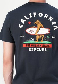 Marine t-skjorte med grafikk av en bjørn som holder et surfebrett, med teksten "CALIFORNIA", "THE GOLDEN STATE" og "RIP CURL" i sterke farger.