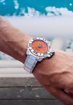 Montre en acier inoxydable avec un cadran orange, des accents noirs et un bracelet argenté. Présente des index horaires audacieux et une lunette unidirectionnelle.