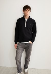 Sweatshirt en polaire noir zippé avec un col haut, porté par-dessus une chemise blanche, associé à un jean gris et des baskets. Texture lisse, coupe décontractée.