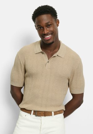Lächelnder Mann mit kurzen lockigen Haaren trägt ein beiges, strukturiertes kurzärmeliges Polohemd und weiße Hosen mit braunem Gürtel und steht vor einem weißen Hintergrund.