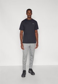 Μαύρο μπλουζάκι Nike με κοντά μανίκια, ανοιχτογκρι φόρμα, και μαύρα αθλητικά παπούτσια, στέκεται σε απλό φόντο. Μινιμαλιστικός σχεδιασμός με διακριτικό λογότυπο.