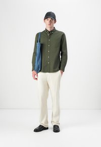 Un giovane uomo in piedi indossa un cappello nero, una camicia verde oliva con bottoni, pantaloni crema, scarpe nere e porta una borsa a tracolla blu sulla spalla, contro uno sfondo bianco.
