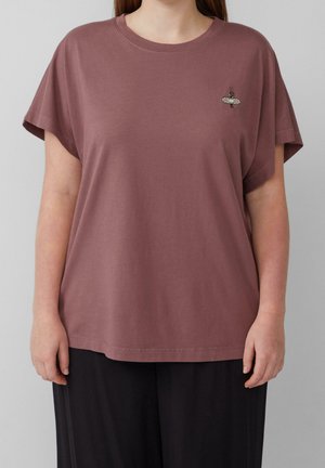 Femme portant un t-shirt mauve ample avec un petit motif de pansement et de fleur sur la poitrine gauche et un pantalon noir, debout devant un fond gris.