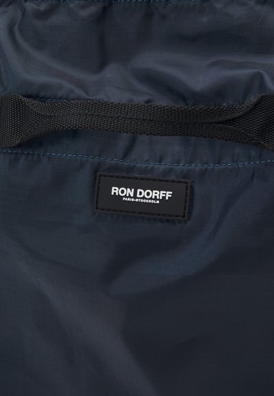 Jūras spēku zila neilona soma ar strukturētu virsmu, melnu austu rokturi un taisnstūrveida etiķeti ar baltu tekstu: "RON DORFF PARIS-STOCKHOLM."