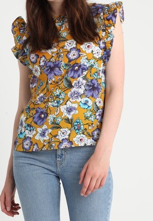 Květinová halenka v hořčicově žluté barvě s krátkými volánovými rukávy; zdobená velkými fialovými a bílými květy se zelenými listy, měkká bavlněná textura.