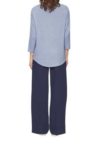 Persoon in een lichtblauwe langslavige top en brede marineblauwe broek, staande met de rug naar de kijker tegen een witte achtergrond.