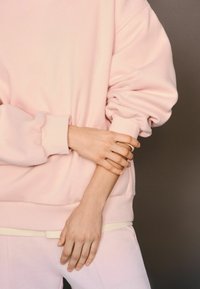 Felpa rosa chiaro con maniche ampie e polsini risvoltati, dalla texture morbida, abbinata a pantaloni coordinati, con un dettaglio sottile di vita gialla.