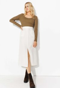 Haut à manches longues vert olive avec un col festonné, associé à une jupe midi blanche avec une fente à l'avant et deux poches plaquées. Bottes à cheville marron.