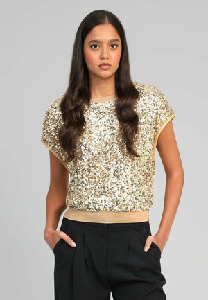 Jeune femme aux longs cheveux foncés portant un haut à manches courtes orné de sequins dorés et un pantalon noir, les mains dans les poches, fond gris neutre.