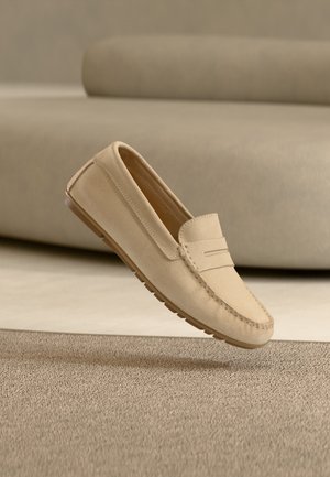 Mocassin en daim beige avec un bout mocassin cousu et une semelle en caoutchouc, flottant au-dessus d'un tapis texturé dans une pièce aux tons neutres.