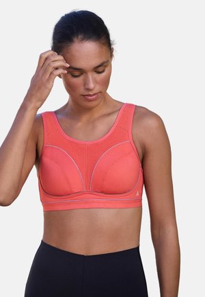 Kvinde iført koralfarvet sports-bh og sorte leggings, der kigger ned med den ene hånd, der rører ved hendes hår.