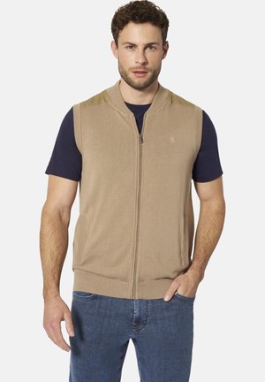 Mann mit kurzen lockigen Haaren, der eine beige Reißverschlussweste über einem dunkelblauen T-Shirt und blauen Jeans trägt, steht mit einer Hand in der Tasche.