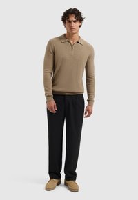 Beige gebreid polo sweater met lange mouwen en een ritskraag, gecombineerd met zwarte losse broek en lichtbruine instap schoenen.