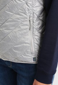 Gray quilted vest with a zipper pocket, featuring a smooth texture and contrasting navy sleeve. Shows partial view of denim jeans.