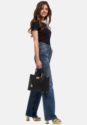 Borsa nera con dettagli in oro, forma rettangolare, superficie testurizzata e un ciondolo attaccato. Il modello indossa una camicia nera, jeans a vita alta e sandali con plateau dorati.