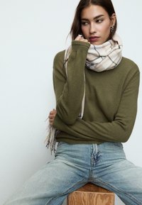 Oliwkowy sweter z dzianiny w połączeniu z jasnym szalikiem w kratę, noszony z jasnoniebieskimi spodniami typu high-waist, siedzący na drewnianym bloczku.