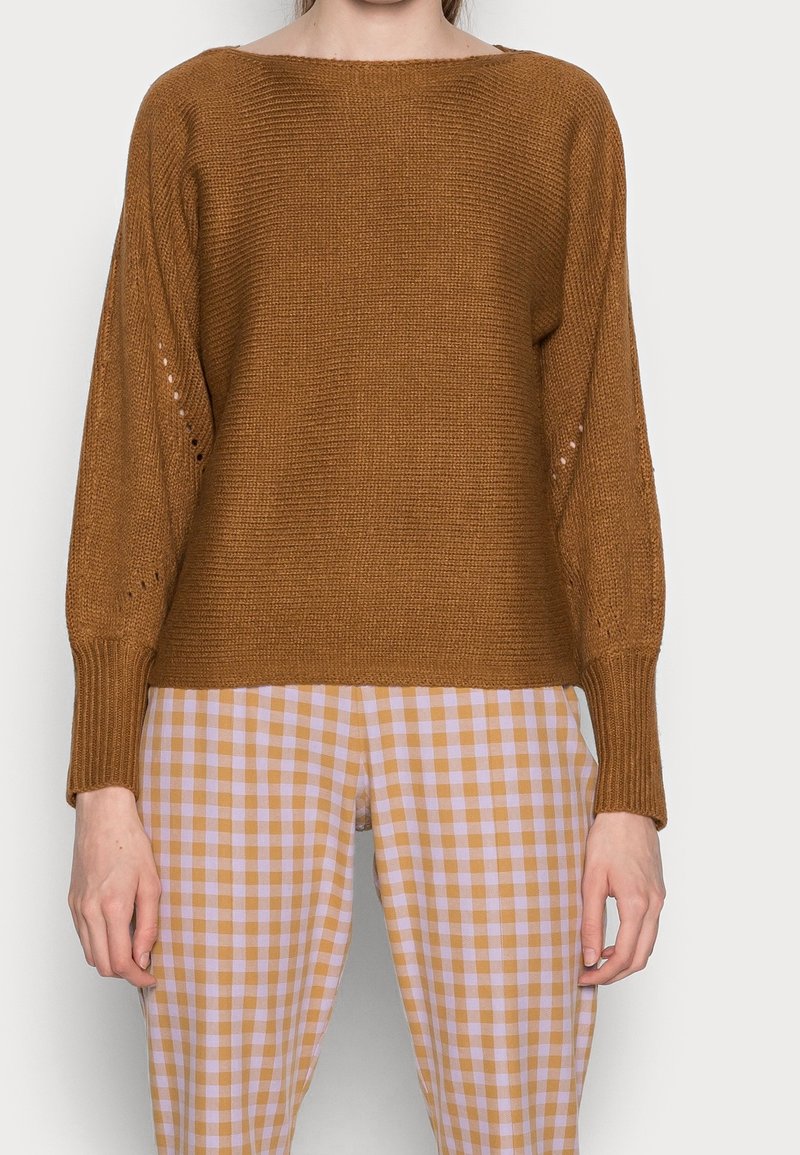 Femme portant un pull en maille marron avec de longues manches bouffantes et un pantalon à carreaux beige et blanc, debout devant un fond uni.