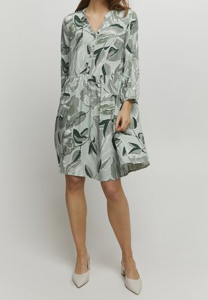 Kobieta w sukience za kolano z zielono-szarym liściastym wzorem, z długimi rękawami, w białych butach na obcasie z czubkiem w szpic, stoi na tle białego tła.