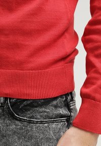 Pull en tricot rouge avec des poignets et un ourlet côtelés, associé à un jean en denim gris foncé, affichant une coupe classique et une texture douce.