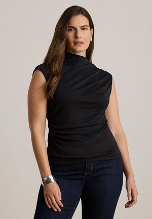 Femme aux longs cheveux bruns portant un haut noir sans manches, un jean foncé, un bracelet en argent et des boucles d'oreilles en argent en forme de créoles, debout devant un fond uni.