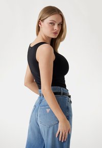 Top nero senza maniche abbinato a jeans in denim blu a vita alta. I jeans presentano una toppa con logo triangolare sulla tasca posteriore.