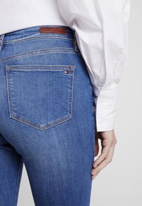 Jeans de mezclilla azul con un diseño slim-fit, que cuentan con un bolsillo trasero y una etiqueta de cuero marrón con la inscripción "TOMMY HILFIGER". La costura es de color amarillo dorado.