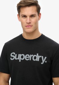 Sort bomulds t-shirt med et stort gråt "Superdry." logo på brystet. Rund hals og korte ærmer. Glat tekstur.