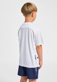 Vit kortärmad idrottströja med en slät textur, med krage och en logotyp tryckt i svart på sidan. Bärs med marinblå shorts.